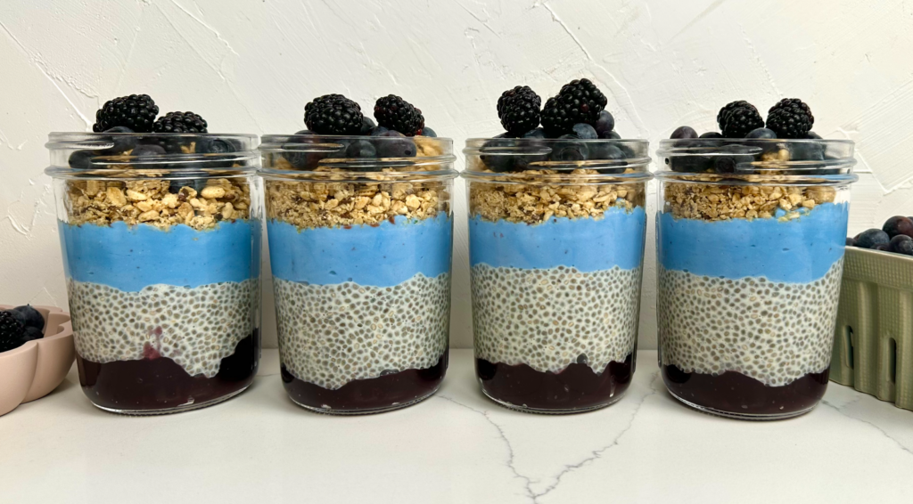 row of 4 meal prep containers showing the layers of blueberry jam, vanilla chia pudding, blueberry yogurt and topped with granola and fresh berries